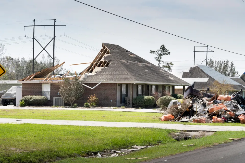 Exterior side view of a single-story brick home missing back half of roof and a debris pile in the front yard.